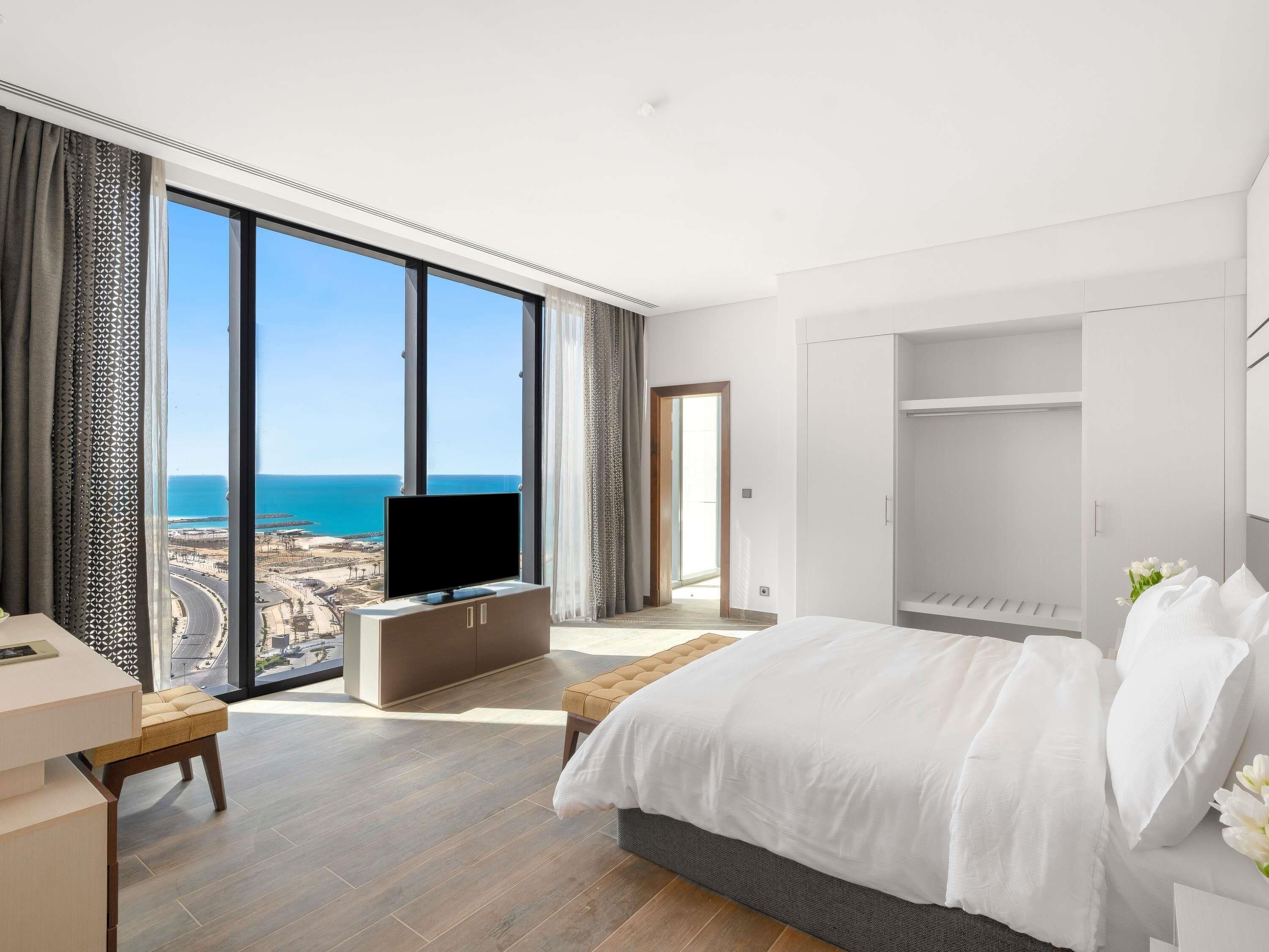 A modern hotel room with a large window, a bed, a TV, a wardrobe, and a desk.