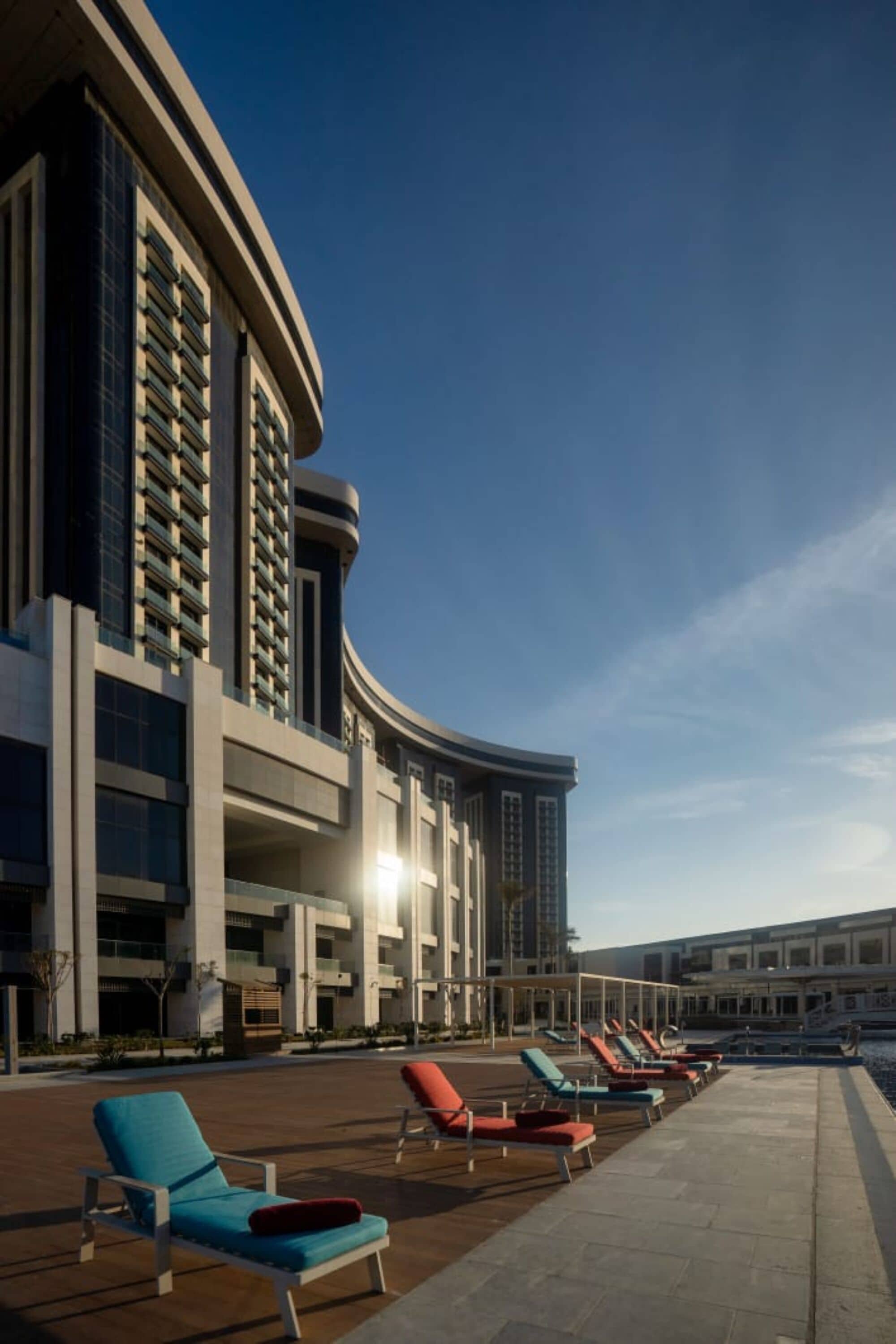A modern building with a curved facade, outdoor seating area with lounge chairs, and a clear sky.