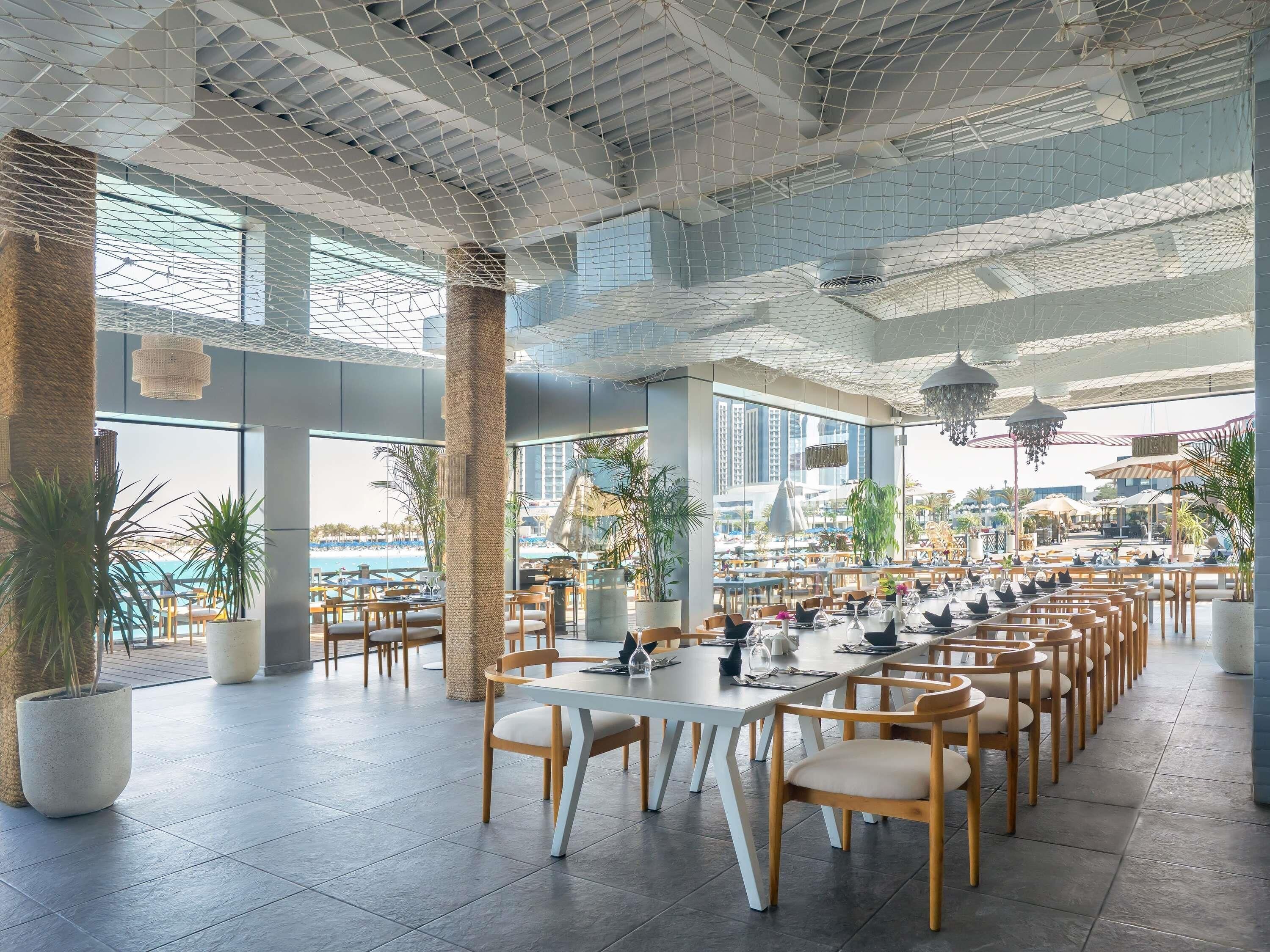 A spacious indoor dining area with multiple tables and chairs, large windows offering views of the cityscape, and a modern ceiling design.