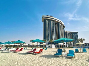 A beach with rows of sun loungers and umbrellas, a large hotel building in the background. - Rixos Premium Alamein (El Alamein)