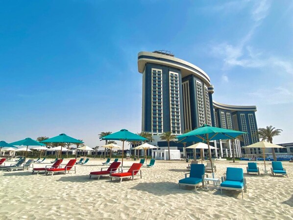 A beach with rows of sun loungers and umbrellas, a large hotel building in the background. - Rixos Premium Alamein (El Alamein)