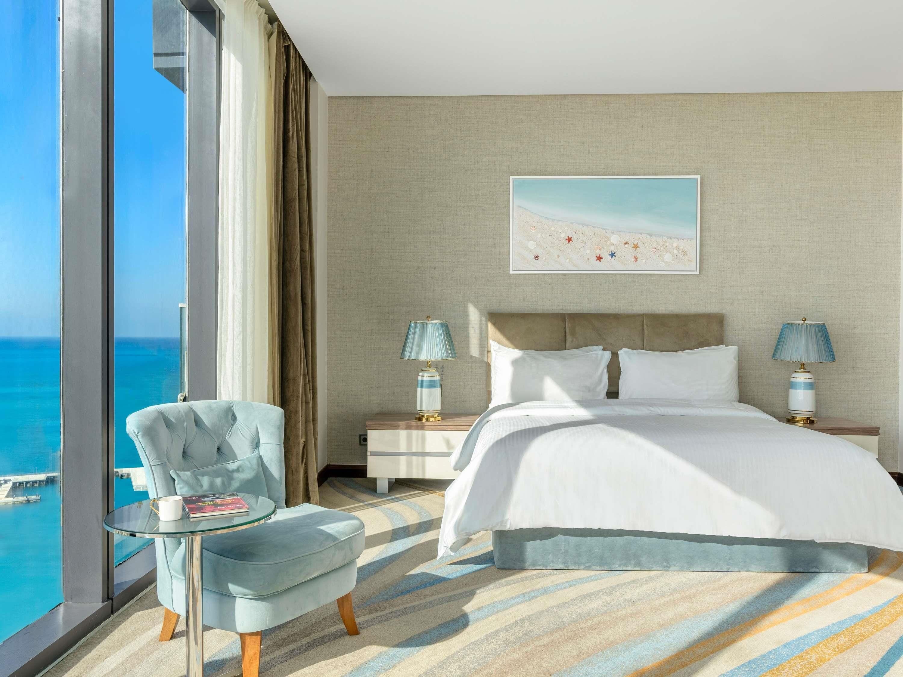 A hotel room with a large bed, two bedside lamps, a chair, a glass table with a book, and a view of the ocean.