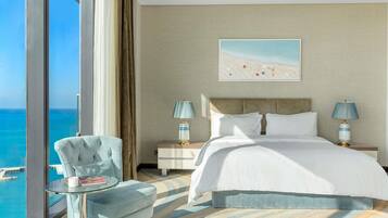 A hotel room with a large bed, two bedside lamps, a chair, a glass table with a book, and a view of the ocean.