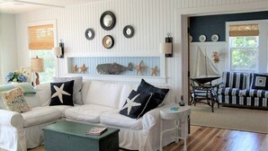 A living room with a white sectional sofa, a small side table, and a coffee table. There are wall-mounted decorations and a window with a view. - 600 Feet to Private Association Cape Cod Bay Beach (2266) (Eastham)