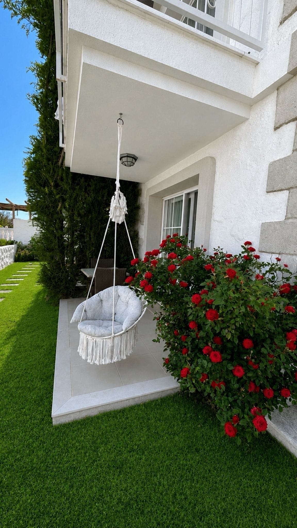 A white building with a hanging chair on a patio, surrounded by green grass and red roses.