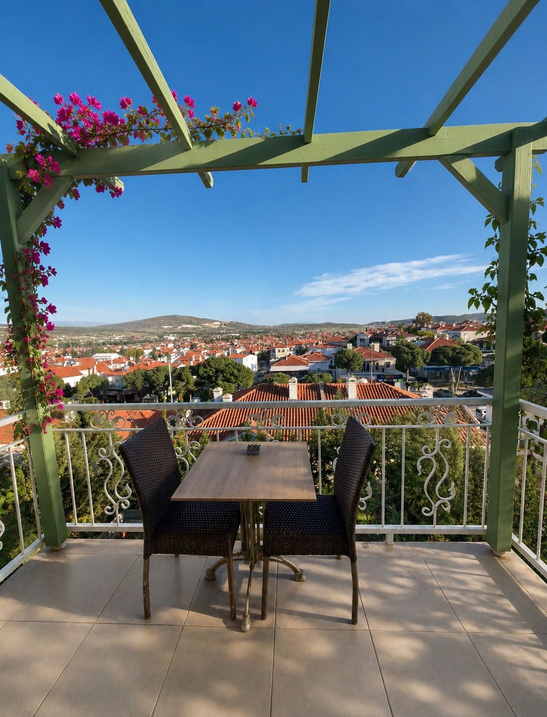 A balcony with a table and chairs, a pergola with flowers, and a view of a town.