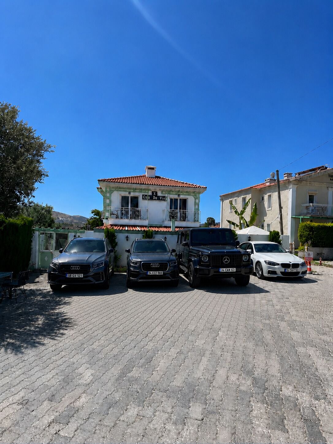 A parking area with three cars parked in front of a building with a tiled roof and a balcony.