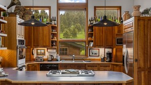 Une cuisine avec des meubles en bois, un îlot central doté de tabourets, une cuisinière à gaz et une grande fenêtre donnant sur une forêt.
