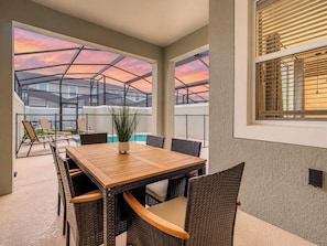 A dining area with a wooden table and wicker chairs, a potted plant, and a view of a swimming pool and buildings. - Sunny Family Escape/ Private Pool & Themed Rooms (Kissimmee)