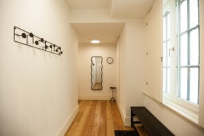 A hallway with wooden flooring, a wall-mounted clock, a mirror, and a window with blinds.