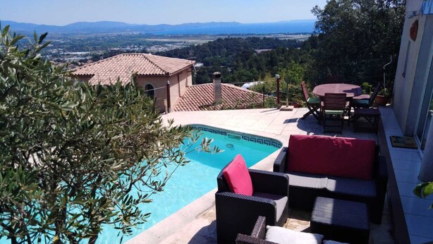A pool area with a view of distant mountains, outdoor furniture, and a building with a tiled roof. - Studio with pool 5 minutes from Hyères beaches. Extraordinary view! (Hyères)