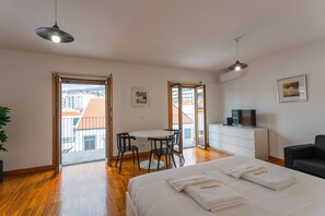 A hotel room with a bed, a desk, chairs, a TV, and a balcony with a view of buildings.