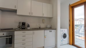 Une cuisine compacte avec des meubles blancs, un four encastré, une machine à café, un lave-linge et une vue dégagée sur l’extérieur grâce à une grande fenêtre.