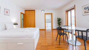 Une chambre d’hôtel avec un lit, un bureau, des chaises, une plante en pot et une vue sur la cuisine par une porte ouverte.