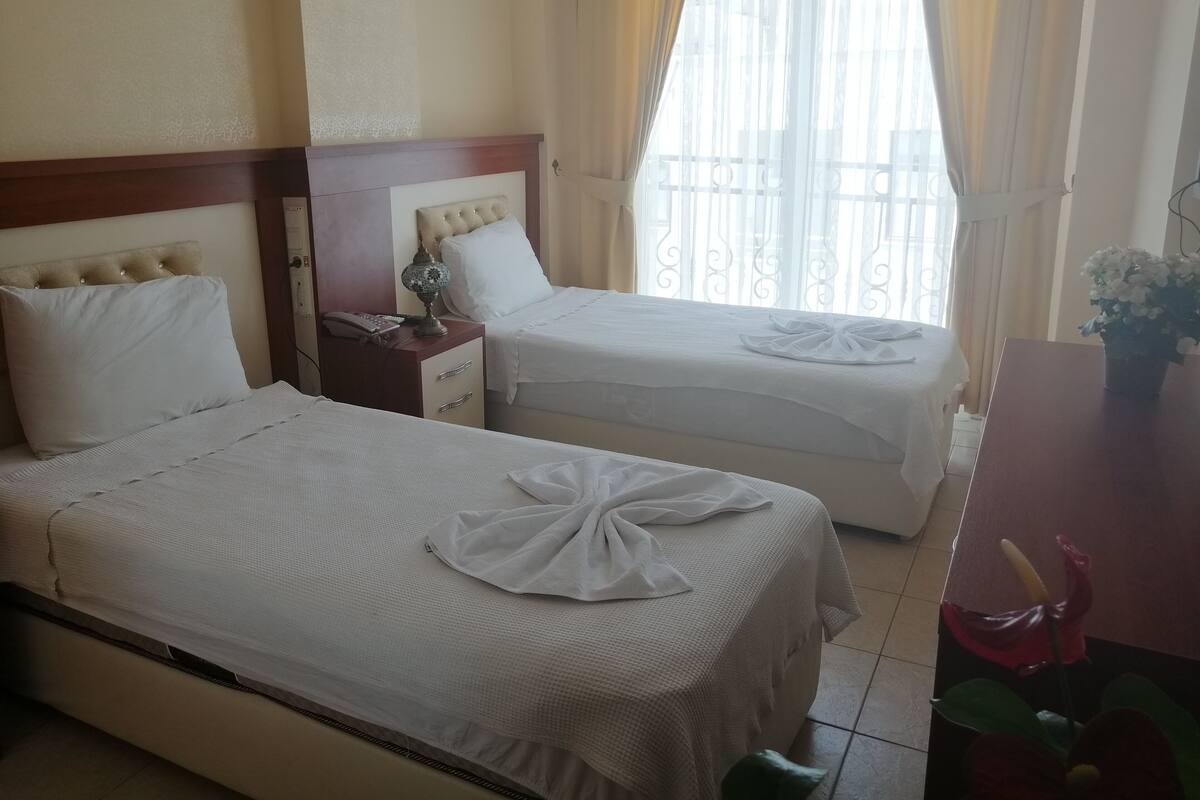 A hotel room with two beds, a wooden headboard, a bedside table with a lamp, and a window with curtains.