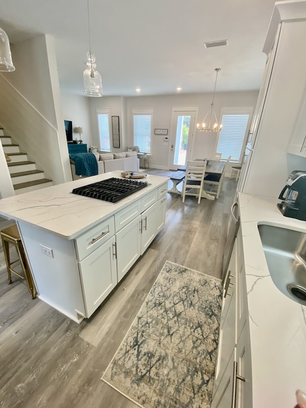 A modern kitchen with white cabinetry, a central island with a stove, and a dining area with a table and chairs. - Beach home w/ 6 seater golf cart, walk to the beach/pool/fast internet (Santa Rosa Beach)