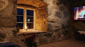 Ein Zimmer mit Steinwänden, einem Fenster und einer kleinen Bank. Auf dem Fernseher ist ein Kaminfeuer zu sehen.