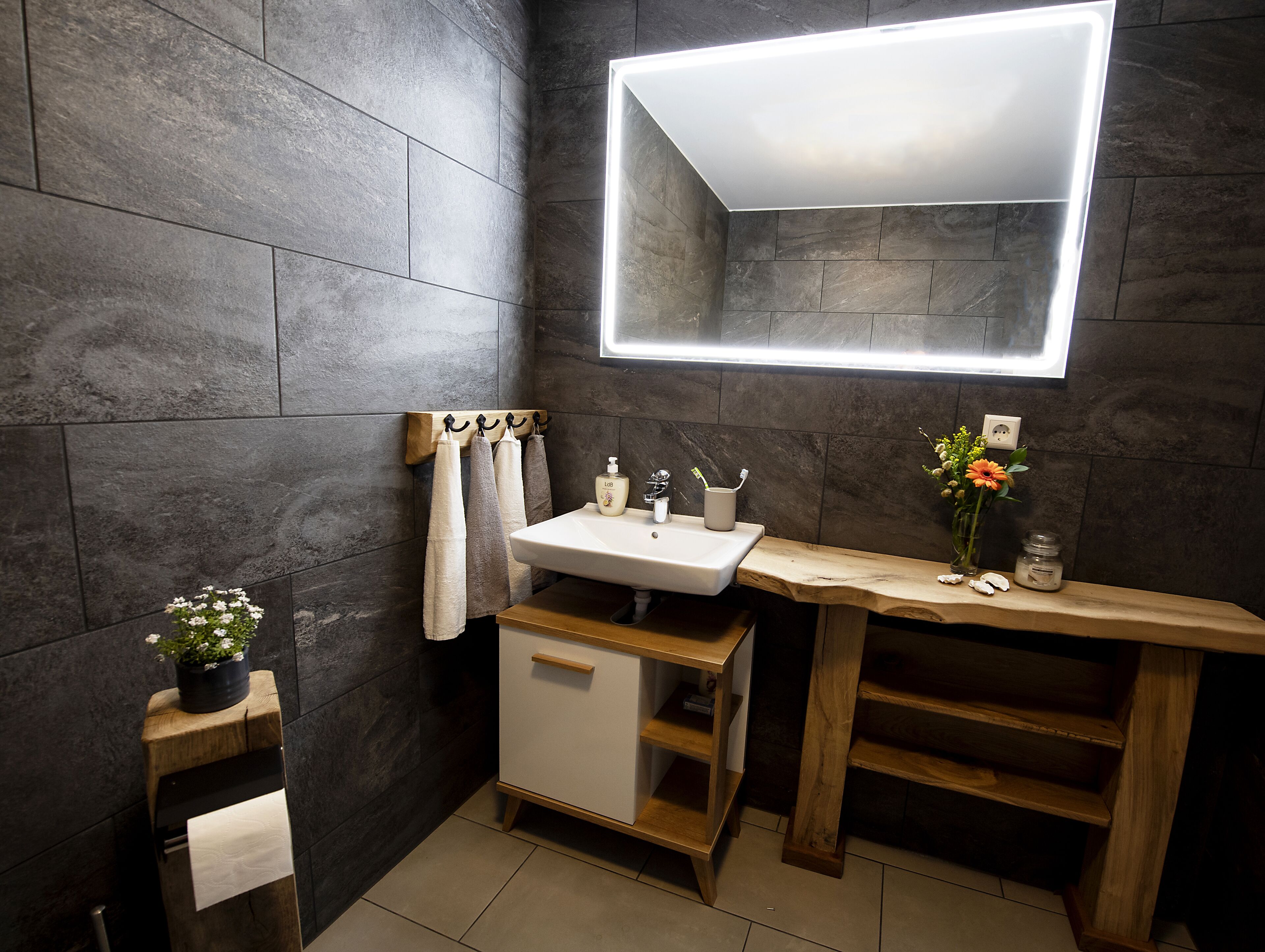 A modern bathroom with a large mirror, dark tiled walls, a wooden vanity, and a small plant.