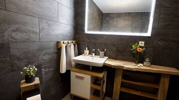 A modern bathroom with a large mirror, dark tiled walls, a wooden vanity, and a small plant.