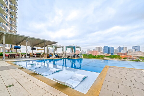 A rooftop pool with lounge chairs and a covered seating area, overlooking a city skyline.