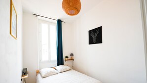 A bedroom with a bed, a window with curtains, a wall-mounted lamp, a framed picture, and a small shelf with a plant. - A break somewhere! (Marseille)