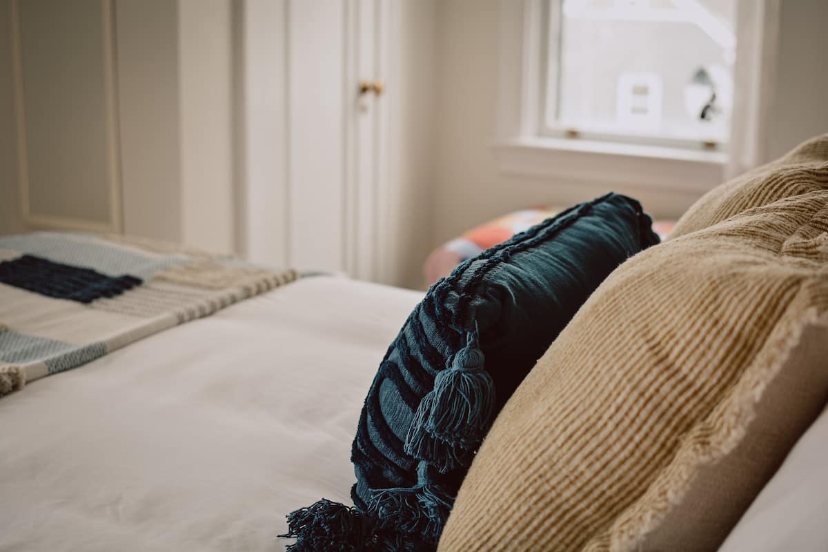 Una cama con una manta color beige y un almohadón azul oscuro, una pared blanca y una ventana con vistas a una casa.