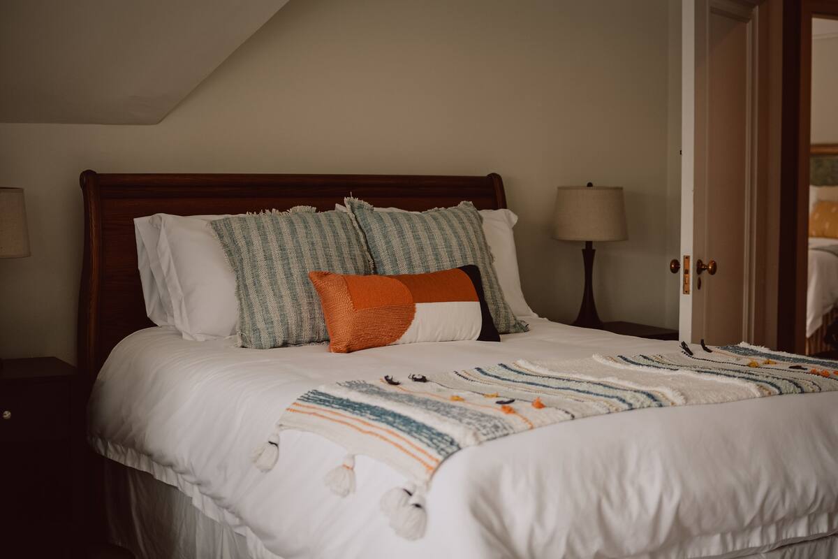 Una cama bien tendida con un edredón a rayas y varias almohadas, una lámpara de noche y un cabecero de madera.