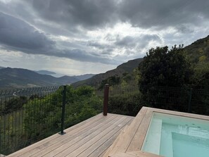 Une terrasse en bois avec une piscine, offrant une vue sur un paysage montagneux, agrémentée d’une clôture et de verdure.