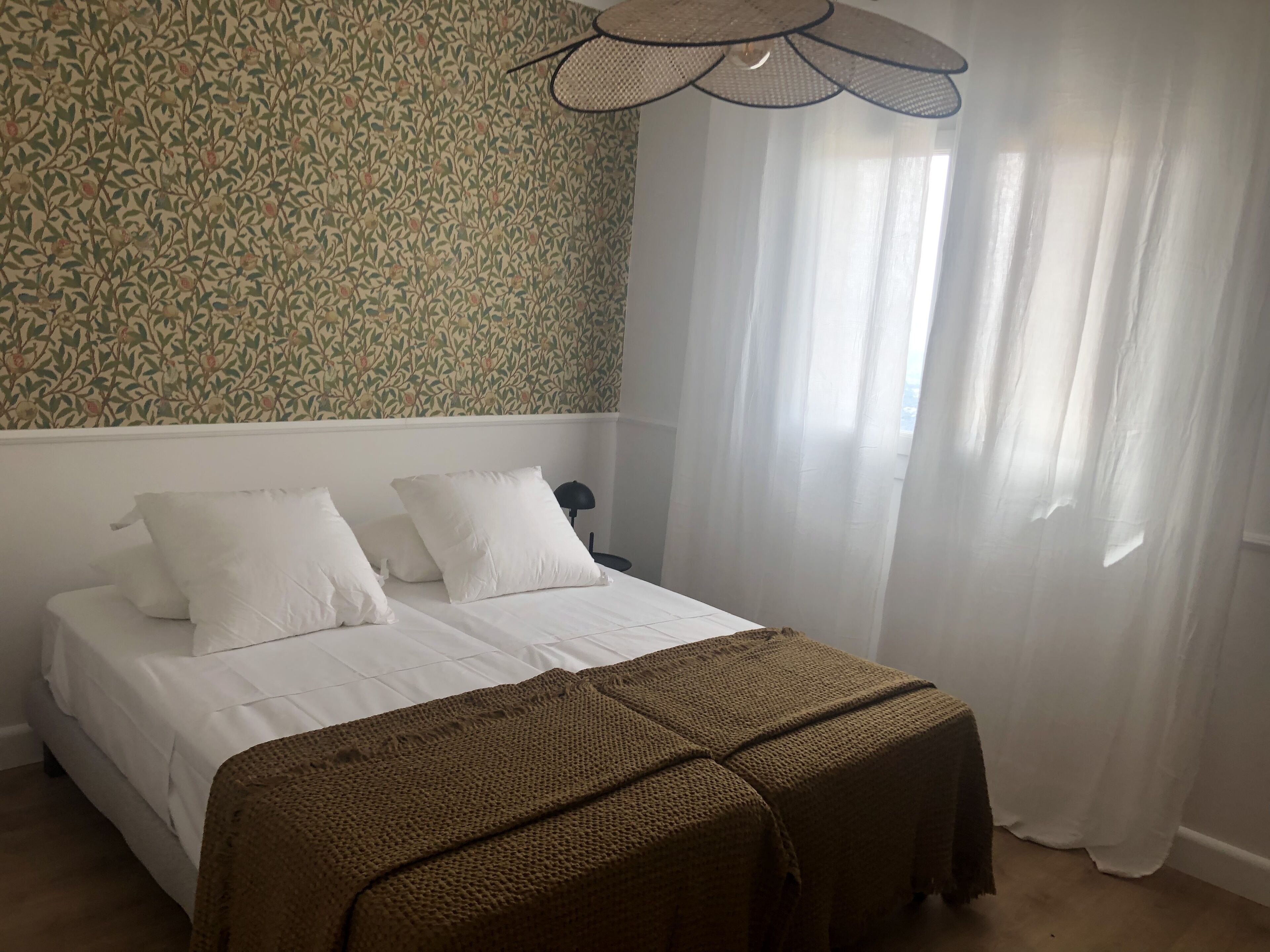 A bedroom with a bed, white bedding, a brown blanket, a patterned wallpaper, and sheer curtains.
