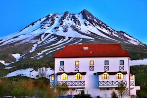 Un edificio de dos pisos con techo rojo y numerosas ventanas, frente a una montaña nevada.