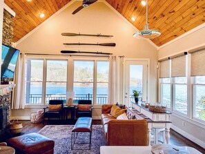 Relax in this spacious living room with a high ceiling, featuring a sofa, bench, and table with a lamp. Enjoy views of the water and forest from the two windows.