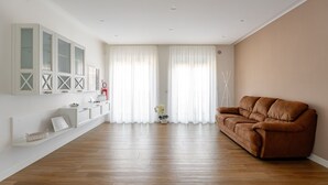 Amplia sala de estar con un sofá marrón, muebles blancos y un gran ventanal con cortinas traslúcidas.