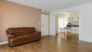 Amplio salón con sofá de cuero marrón, suelo de madera y vistas a una cocina moderna con armarios blancos y zona de comedor.