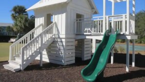 Ein Spielplatz mit einer grünen Rutsche und einem weißen Spielhäuschen mit Treppe.