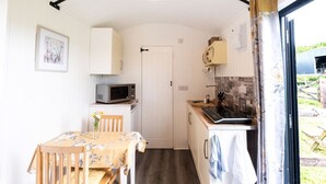 A compact kitchen with white cabinets, a microwave, and a stove. There is a dining table with chairs and a small window with curtains. - Shepherd's hut with hot tub (Bodmin)