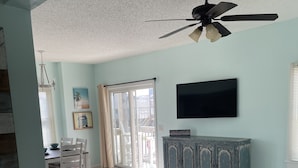 Ein geräumiges Wohnzimmer mit Deckenventilator, Fernseher, Essbereich mit Stühlen, einem Schrank und Blick durch eine Glasschiebetür ins Freie.