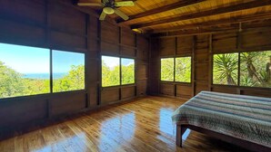 Habitación con piso de madera, una cama, un ventilador de techo y amplias ventanas que dan a vistas de árboles y un paisaje lejano.