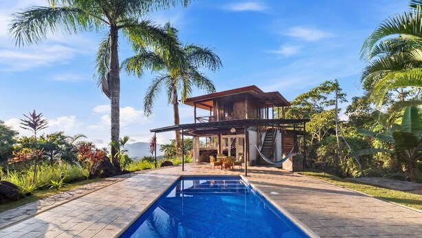Una piscina con cubierta de madera, un edificio de dos pisos con techo rojo y vegetación tropical.