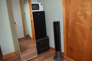 Quarto compacto com piso de madeira, porta branca, geladeira preta e uma coluna de som preta alta.