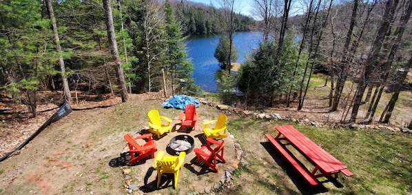 An outdoor seating area with Adirondack chairs and a picnic table, surrounded by trees and a lake.