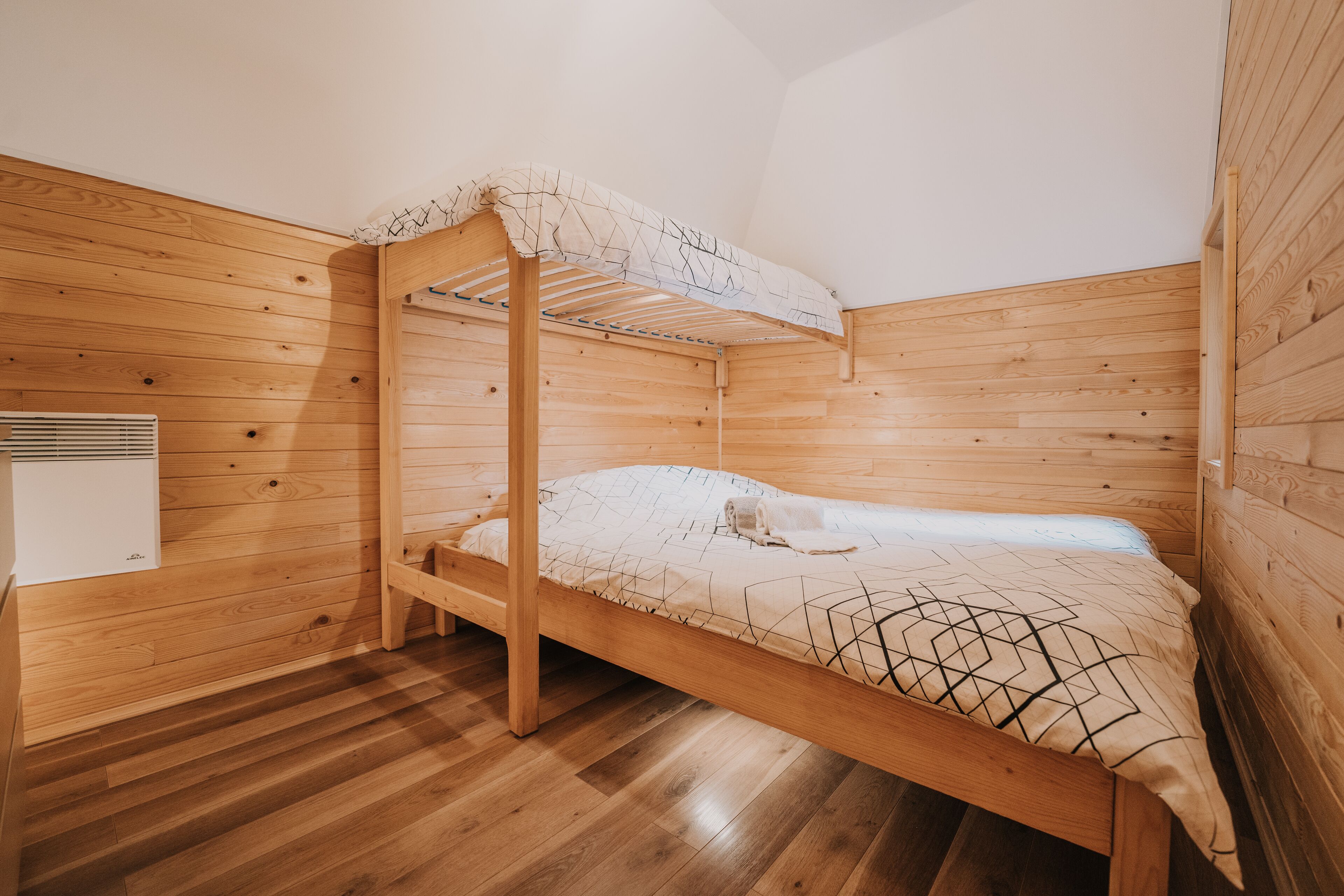 Ein Holzbett mit zwei Etagen, eine Matratze und Kissen, ein Holzboden und eine weiße Wand mit Lüftungsgitter.