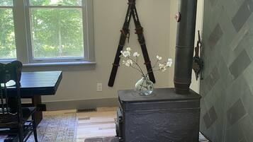 Een kamer met een groot raam, een rustieke houtkachel, een eettafel met stoelen, en een decoratieve glazen vaas met bloemen.