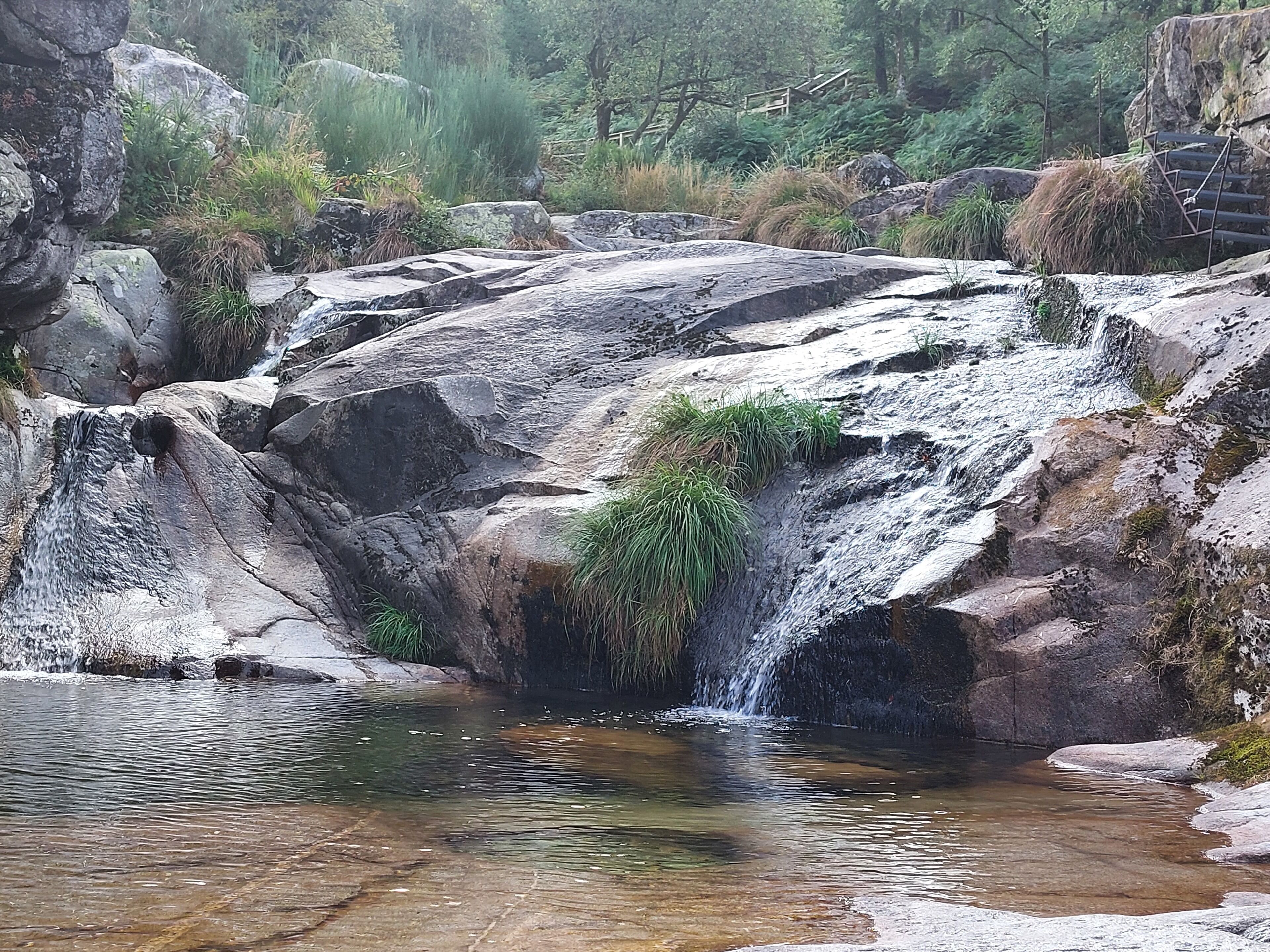 A natural waterfall cascading over rocks into a clear pool, surrounded by greenery and a wooden bridge.