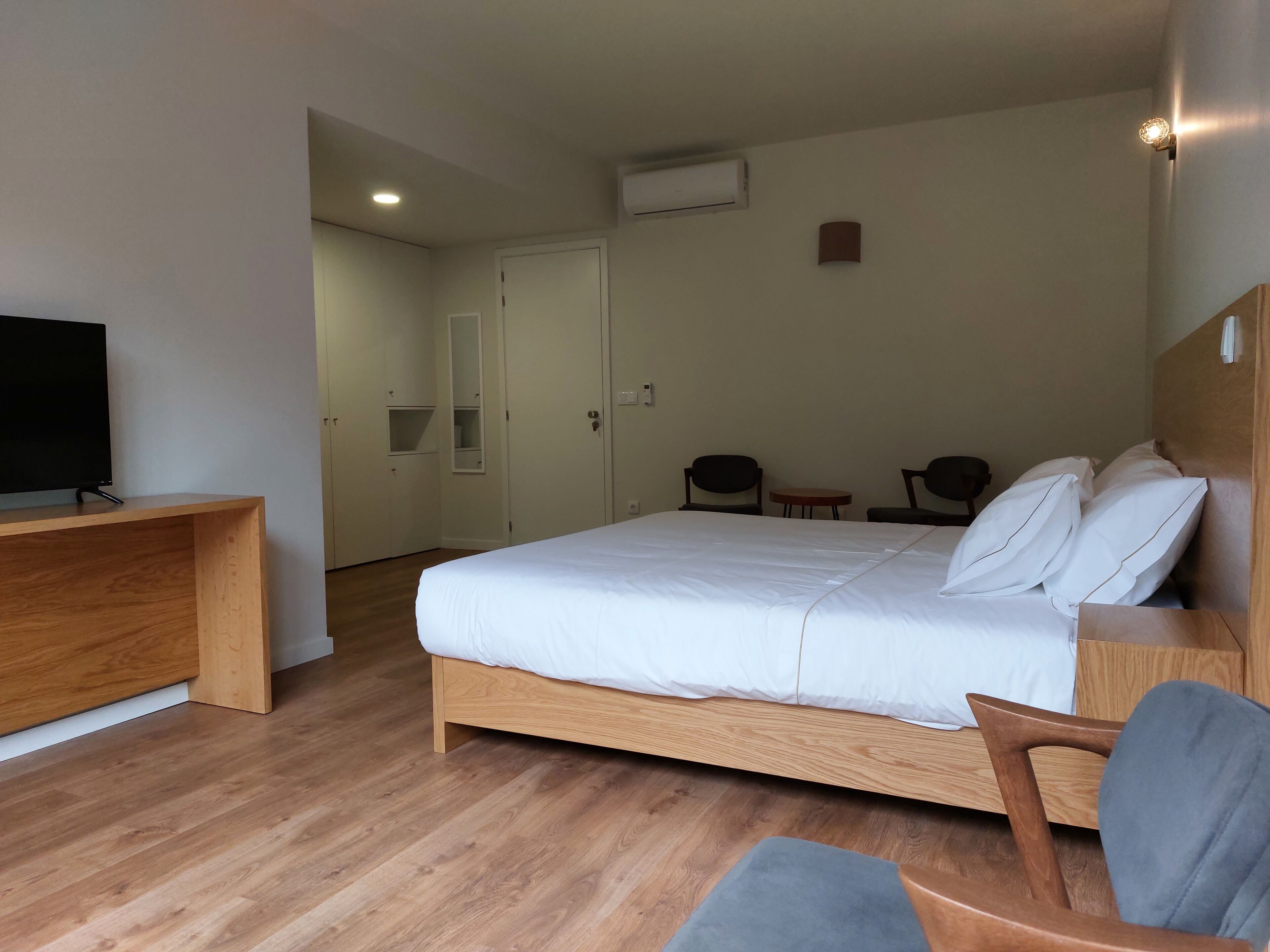 Quarto de hotel com cama de madeira, televisão sobre suporte de madeira e uma pequena mesa com cadeiras.