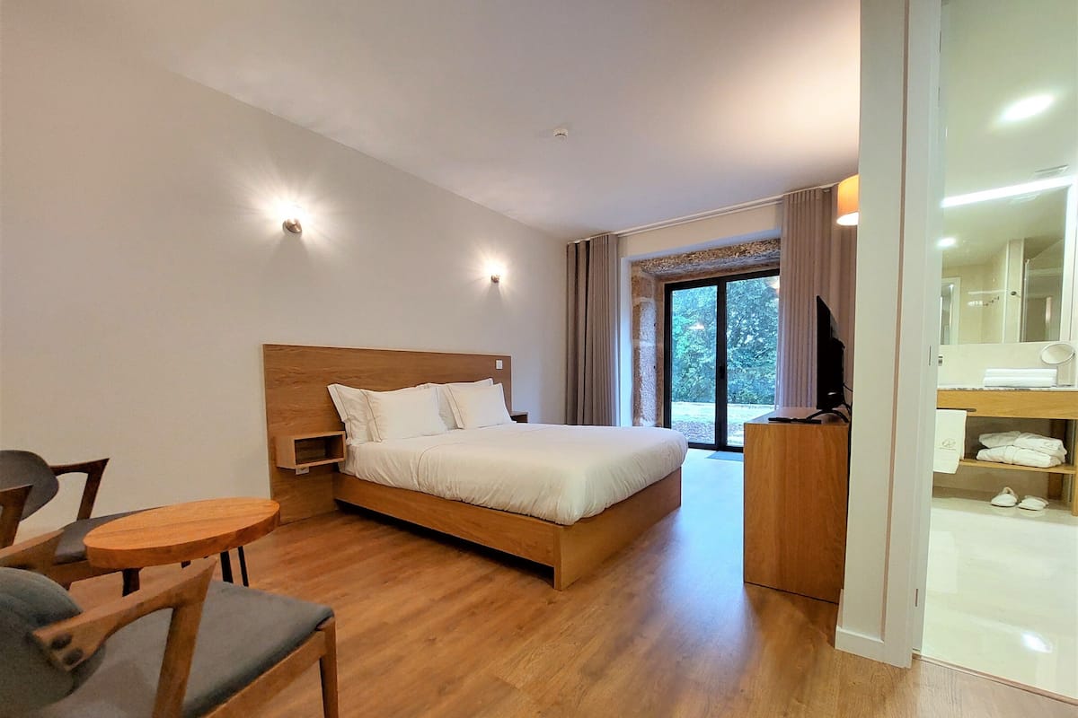 A hotel room with a wooden bed, a small wooden table with a chair, a bathroom with a mirror and towel rack, and a view of trees through the window.