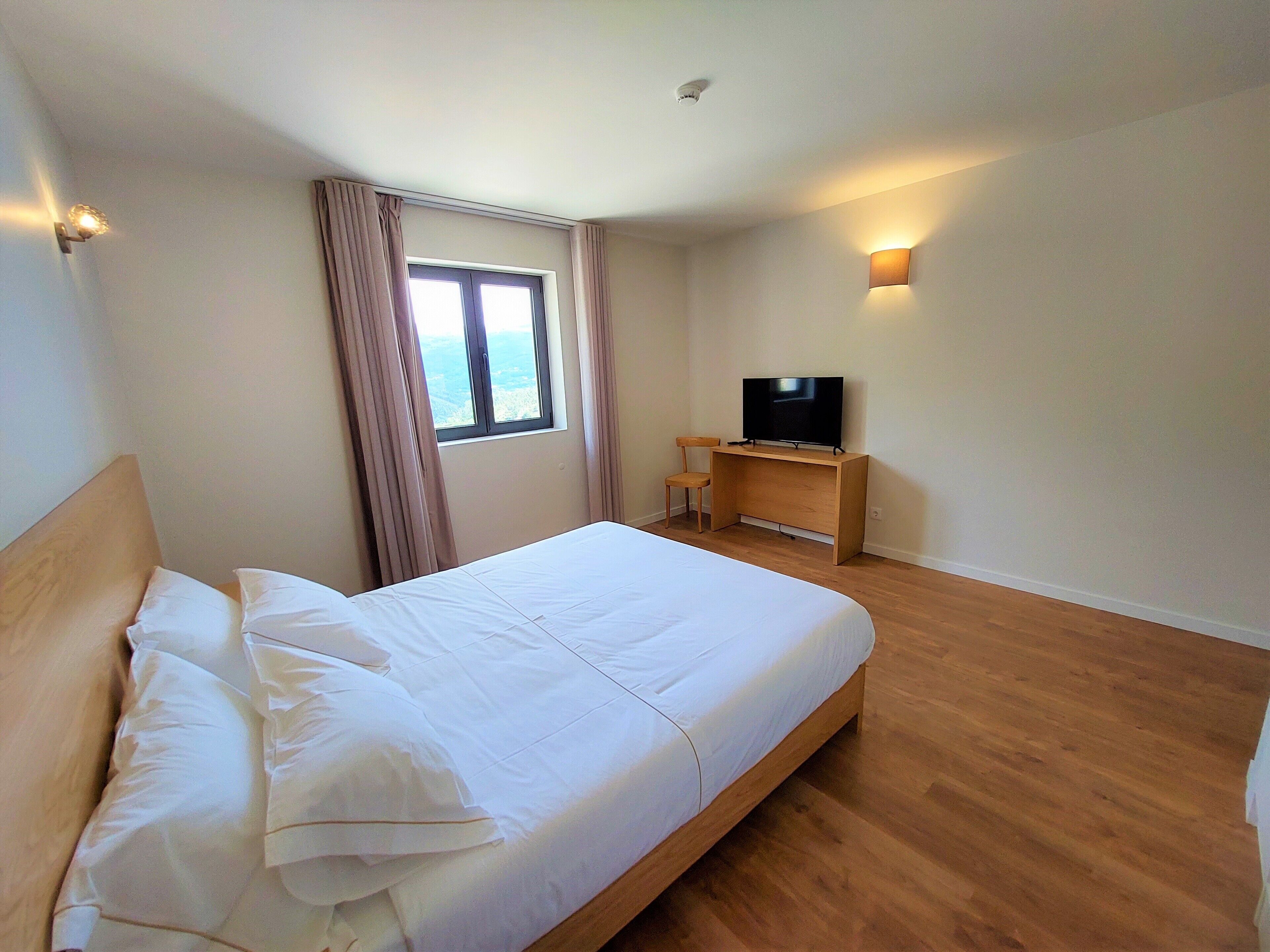 A hotel room with a large bed, a TV on a wooden stand, a window with curtains, and a wall-mounted light.