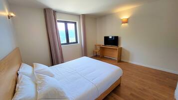 A hotel room with a large bed, a TV on a wooden stand, a window with curtains, and a wall-mounted light.