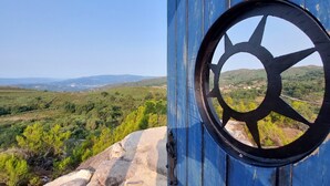 A view through a blue metal gate with a circular window, overlooking a green landscape with hills and a clear sky. - Quinta da Roda - Nature and Wellness (Vila Verde)