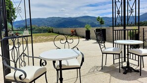 An outdoor patio with round tables and chairs, a view of distant mountains, and a decorative metal gate. - Quinta da Roda - Nature and Wellness (Vila Verde)