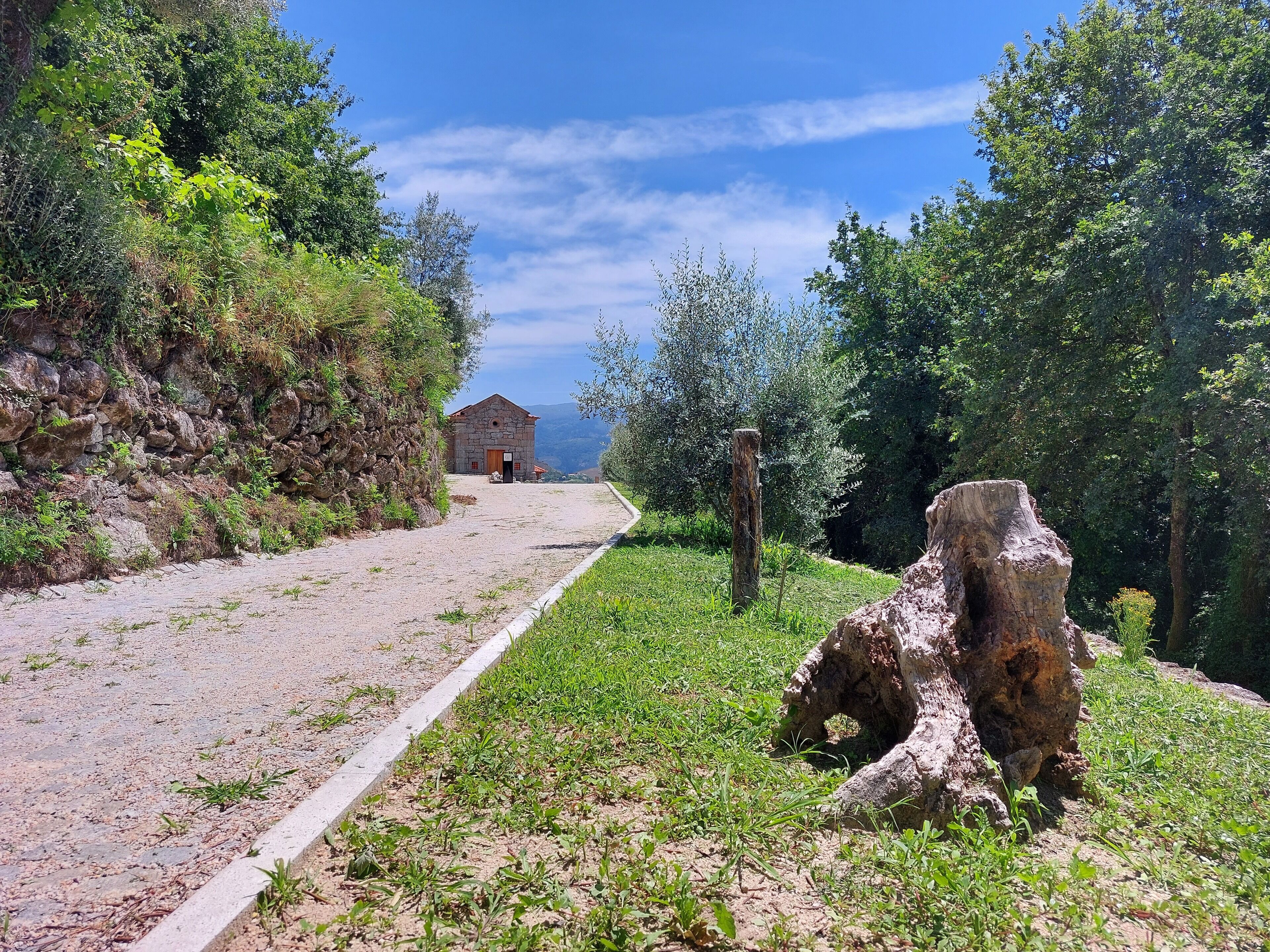 A rural road leading to a stone building with a red roof, surrounded by greenery and a clear sky.